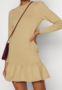 Béžové šaty s dlouhým rukávem a rozšířeným lemem, disponují mírně nařasenými rukávy, kulatým výstřihem a strukturovaným povrchem. Doplněné malou burgundskou kabelkou přes rameno.