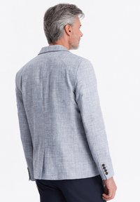 Helles graues Blazer aus strukturiertem Stoff, mitreverskragen und vier dunklen Knöpfen an den Ärmeln. Hat eine taillierte Passform und seitliche Schlitze.