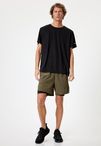Camiseta negra de manga corta combinada con pantalones cortos verde oliva, con un ajuste relajado. El calzado son zapatillas deportivas negras con superficie texturada.