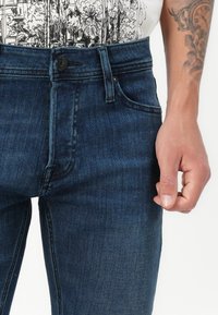 Tuvumā redzama persona, kas ģērbjas tumši zilos džinsos un baltā t-kreklā ar grafisku zīmējumu, parādot tetovētu roku un atslābinātu plaukstu.