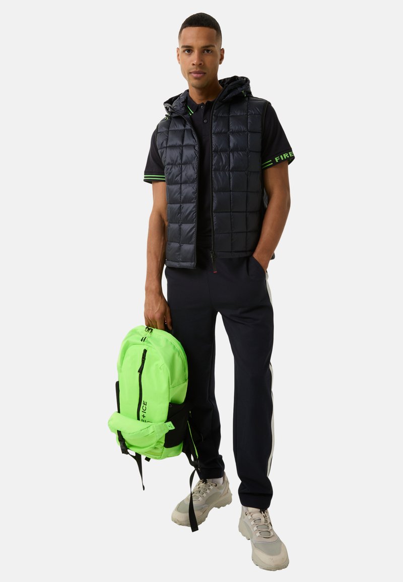 Gepolsterte marineblaue Weste mit Kapuze, schwarzes Poloshirt mit grünen Akzenten, marineblaue Hose und leuchtend grüner Rucksack mit schwarzen Details; graue Sneaker.