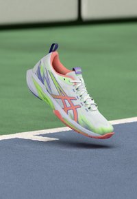 Άσπρο αθλητικό παπούτσι με πράσινες και ροζ πινελιές, επάνω μέρος από δίχτυ, υφή στην σόλα και μοβ ταμπέλα στη φτέρνα, αιωρούμενο πάνω από μια μπλε επιφάνεια γηπέδου.