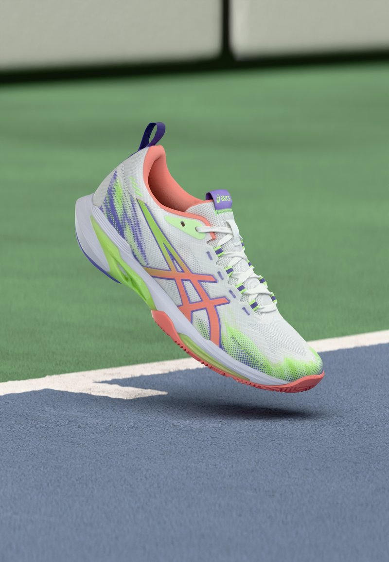 Chaussure de sport blanche avec accents verts et roses, tige en mesh, semelle texturée et languette violette au talon, flottant au-dessus d'une surface de terrain bleue.