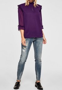 Blusa in chiffon viola con maniche a volant, abbinata a jeans skinny blu sbiaditi con dettagli strappati e scarpe nere in vernice.