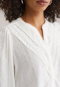 Blouse blanche en tissu texturé, présentant un devant plissé, des détails en dentelle le long de l'encolure et des manches longues bouffantes.