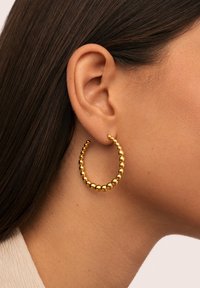 Gros plan sur l'oreille d'une femme avec de longs cheveux noirs portant une boucle d'oreille en or faite de petites perles rondes.