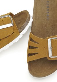 Sandales en daim marron avec semelles en caoutchouc blanc, boucles en argent et détails découpés sur les sangles. Semelle intérieure texturée visible.