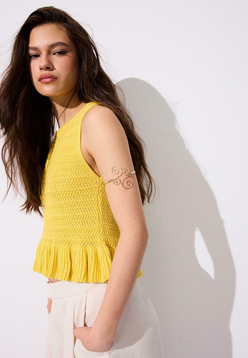 Jeune femme aux longs cheveux bruns porte un top tricoté jaune sans manches et un pantalon clair, debout contre un mur blanc avec une ombre.