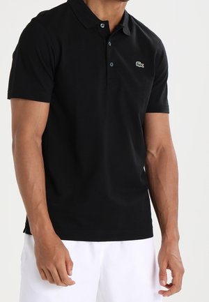 Mann trägt ein schwarzes kurzärmliges Polohemd mit kleinem Krokodil-Logo auf der Brust und weiße Shorts.