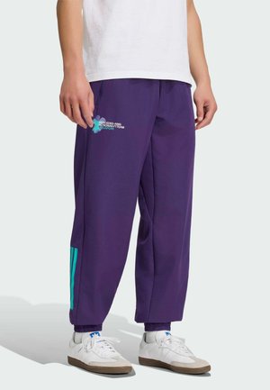 Persona che indossa pantaloni della tuta viola con strisce turchesi e testo "Mercedes-AMG Petronas F1 Team Singapore", sneakers bianche e una camicia bianca.