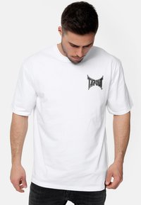 Bílý bavlněný tri�čko s černým grafickým logo "TAPOUT" na levé hrudi. Klasický kulatý výstřih a krátké rukávy.