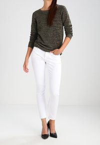 Olivgrön texturerad tröja med långa ärmar, kombinerad med figurnära vita jeans och svarta stilettklackar, mot en enfärgad bakgrund.