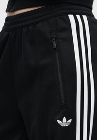 Close-up van zwarte Adidas broek met witte drie strepen, ritssluiting aan zak en logo bij de heup, gedragen met een zwart shirt.