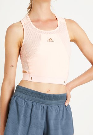 Vrouw die een lichtroze Adidas sportbeha en losse blauwe sportshorts draagt, staand met één hand op haar heup.