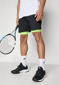 Shorts de sport noirs avec des accents vert néon, associés à des baskets noires et des chaussettes blanches. Tient une raquette de tennis avec un grip à motifs.