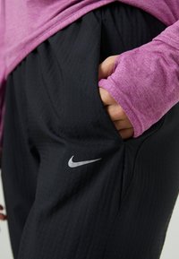 Czarne spodnie sportowe z teksturowanej tkaniny, z boczną kieszenią i małym srebrnym logo Nike na udzie. W połączeniu z różową bluzką z długim rękawem.