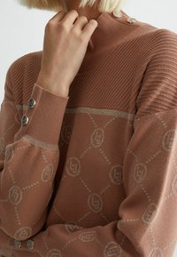 Pull tricoté marron clair avec texture côtelée, motif argenté décoratif et boutons décoratifs sur l'épaule.
