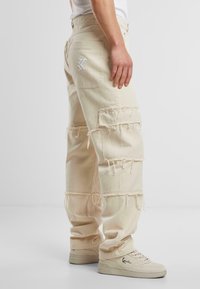 Pantaloni cargo bej cu margini destrămate, două buzunare în față și cusături decorative pe buzunarul din spate. Asortați cu adidași bej.