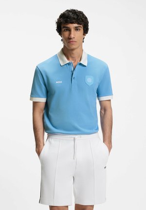 Joven con una camisa polo azul claro con cuello y puños blancos, combinada con pantalones cortos blancos con cordón, de pie con las manos en los bolsillos.