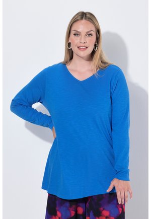 Frau trägt ein hellblaues langärmeliges Oberteil und bunte gemusterte Hosen, steht mit einer Hand in der Hüfte vor neutralem Hintergrund.