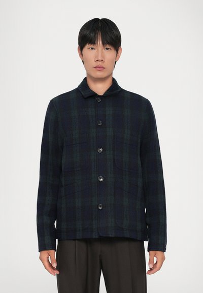 Hombre vestido con una chaqueta de cuadro de botones en tonos azul oscuro y verde, con bolsillos frontales, combinada con pantalones oscuros holgados, de pie frente a un fondo blanco.