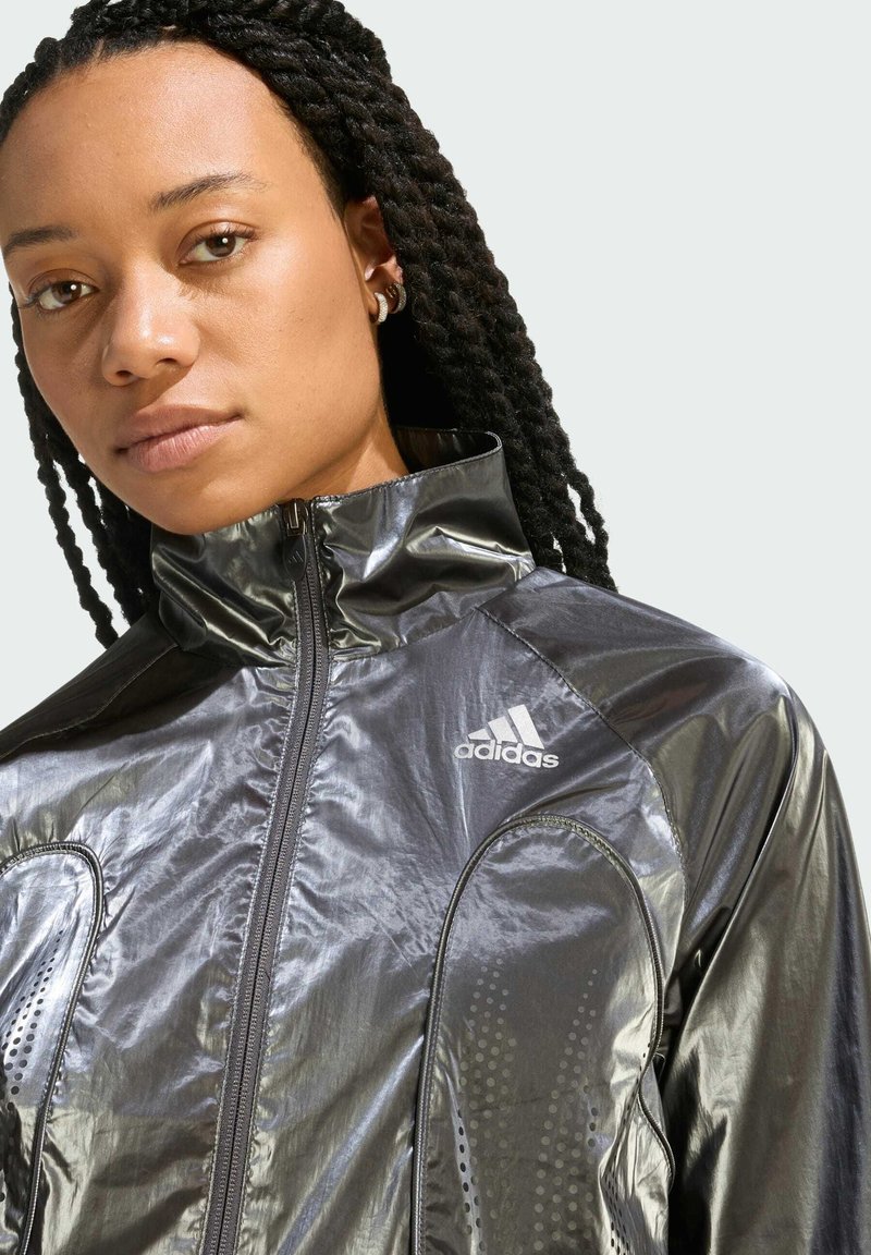 Black, shiny Adidas jacket with high collar, zip closure, and dotted texture accents on sides. Features white logo on chest.
