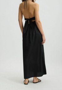 Mustat halterneck-mekko, jossa on avoin selkä ja solmuddetaali. Valoisa kangas ja nilkkapituinen muotoilu, jossa on sileä tekstuuri ja minimaalinen varustus.