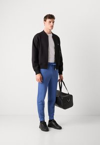 Zwarte bomberjack, grijs t-shirt, blauwe joggers, zwarte sneakers en een zwarte sporttas. Gladde materialen zonder zichtbare patronen of accenten.