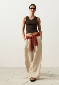 Woman wearing sleeveless dark brown top, wide beige pants, red knit sweater tied around waist, dark sunglasses, and black platform sandals.