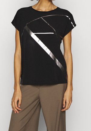 Frau trägt ein schwarzes kurzärmeliges Oberteil mit metallisch glänzenden geometrischen diagonalen Streifen und lockere taupefarbene Hose, steht vor neutralem Hintergrund.