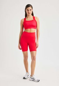 Una mujer de pie, vistiendo un sujetador deportivo rojo y unos pantalones cortos ciclistas de cintura alta a juego, zapatillas de correr blancas, fondo neutro.