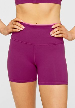 Woman wearing high-waist purple athletic shorts with hands resting on hips against a plain background.
