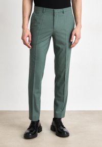 Groene op maat gemaakte broeken van geweven stof, met een rechte pijp en scherpe plooien. Gecombineerd met zwarte veterschoenen.
