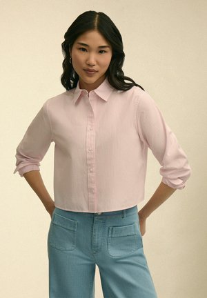 Jonge vrouw die een lichtroze blouse met opgerolde mouwen draagt en een blauw broek met hoge taille en voorzakken.