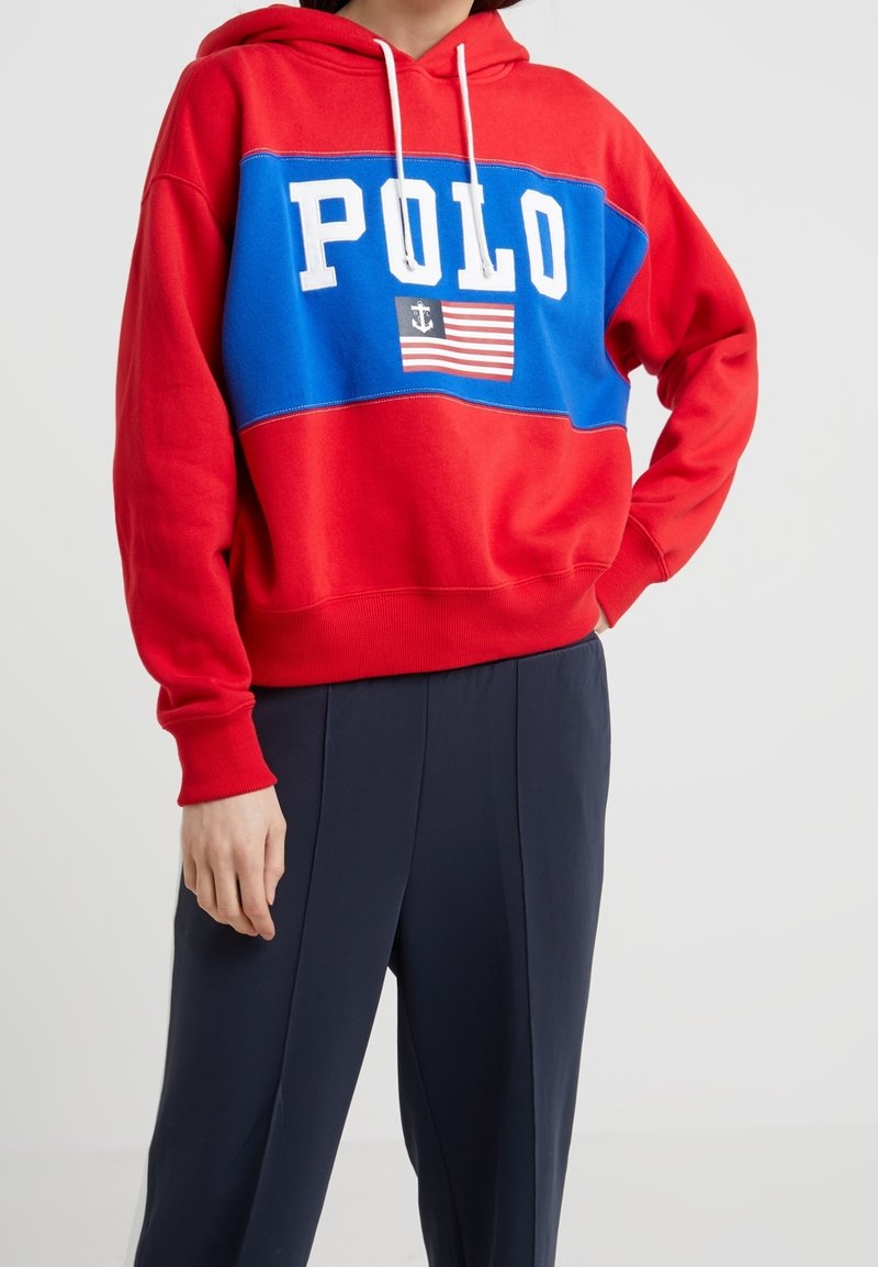 Sweat à capuche rouge et bleu avec le texte "POLO" blanc et un emblème de drapeau, porté avec un pantalon bleu foncé, personne debout avec une main sur la hanche.