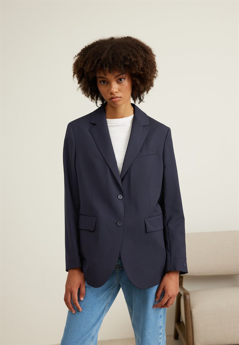 Blazer marine à deux boutons, avec des revers en notch et des poches avant. Coupe ajustée avec une texture lisse, assorti à un t-shirt blanc et à un jeans.