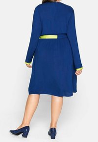 Robe bleu marine à manches longues et taille cintrée, avec une ceinture et des poignets contrastants en vert néon. Associée à des talons à motifs.