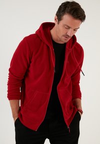 Crvena zip-up dukserica od mekog, teksturiranog materijala s prednjim džepom i podesivim trakama na kapuljači. Nosena preko crne majice.