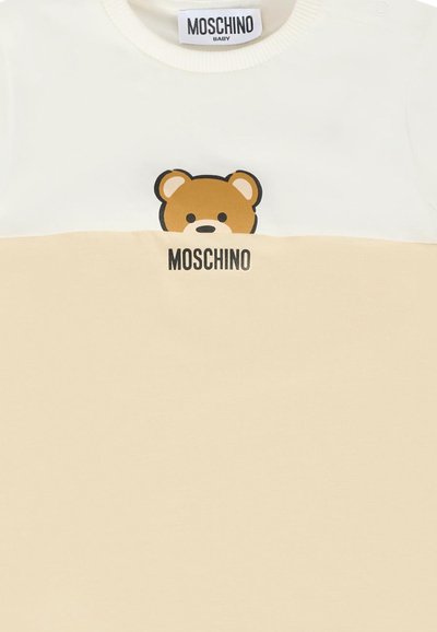 Maglietta per bebè crema e beige con una grafica parziale di un orsetto e il logo "Moschino" stampato sul davanti, sotto il colletto.