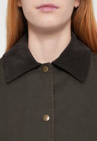 Καφέ μπουφάν με γιακά από κοτλέ, κατασκευασμένο από σκούρο πράσινο ύφασμα, με κλείσιμο με κουμπιά τύπου snap και λεία υφή. Κοντινή λήψη του γιακά και του πάνω μέρους του υφάσματος.