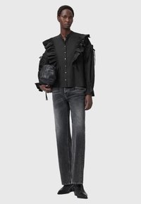 Blusa nera con maniche a ruffles, bottoni frontali, abbinata a jeans grigi e stivali neri alla caviglia. Porta una borsa rotonda nera.