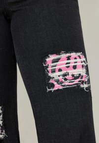 Jeans in denim nero con toppe strappate e un design di faccina sorridente rosa. Il tessuto è leggermente testurizzato con cuciture visibili e sfilacciature.