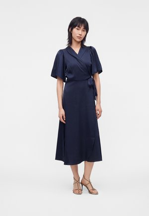 Femme portant une robe portefeuille bleu marine à manches courtes bouffantes et des sandales à talons beige, debout devant un fond blanc uni.