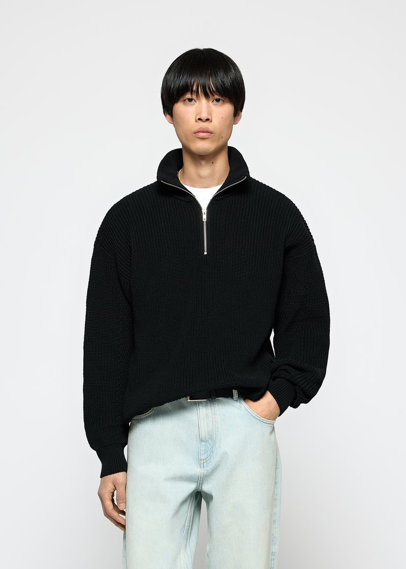 Svart strikket genser med høy krage og kvart glidelås, med ripet struktur og løs passform, kombinert med lyse denimjeans.