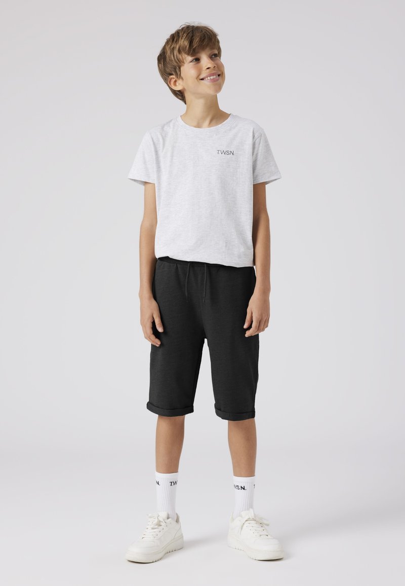 Junge steht lächelnd da, trägt ein hellgraues T-Shirt, schwarze knielange Shorts, weiße Socken und weiße Turnschuhe vor einem schlichten Hintergrund.