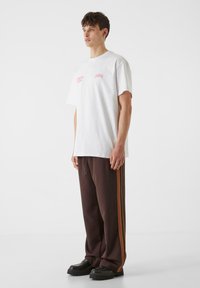Biela oversize tričko s detailom ružového loga, v kombinácii s hnedými teplákmi so bočnými oranžovými pruhmi a čiernymi pevným obuvou.