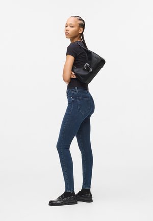 Black handbag with silver hardware, worn on one shoulder; fitted black tee; dark blue skinny jeans; black loafers with studded accents.