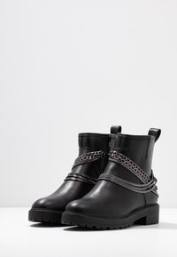 Botas de tobillo de cuero negro con detalle de cadena en la parte superior, suela de goma resistente y tiradores en la parte trasera para facilitar su uso.