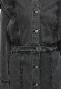 Gros plan sur l'avant d'une veste en denim gris foncé avec des boutons-pression en argent et un détail au niveau de la taille.