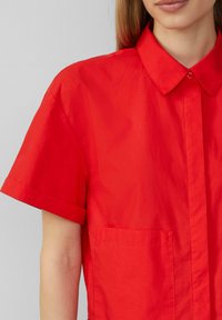 Czerwona koszula z krótkim rękawem z lekkiego materiału, z kołnierzykiem zapinanym na guziki i kieszenią naszywaną po lewej stronie. Gładka tekstura.